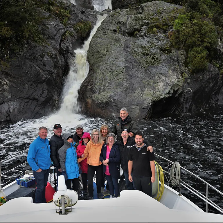 Tour group gathered on the bow of the Southern Secret smiling for the camera with waterfall in the background.